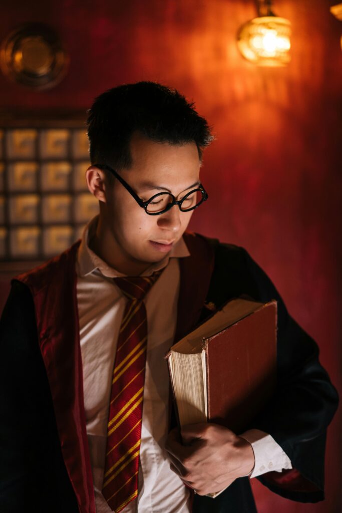 a young asian man with glasses and a jacket and tie holds and immense old book in moddy orange lighting.
